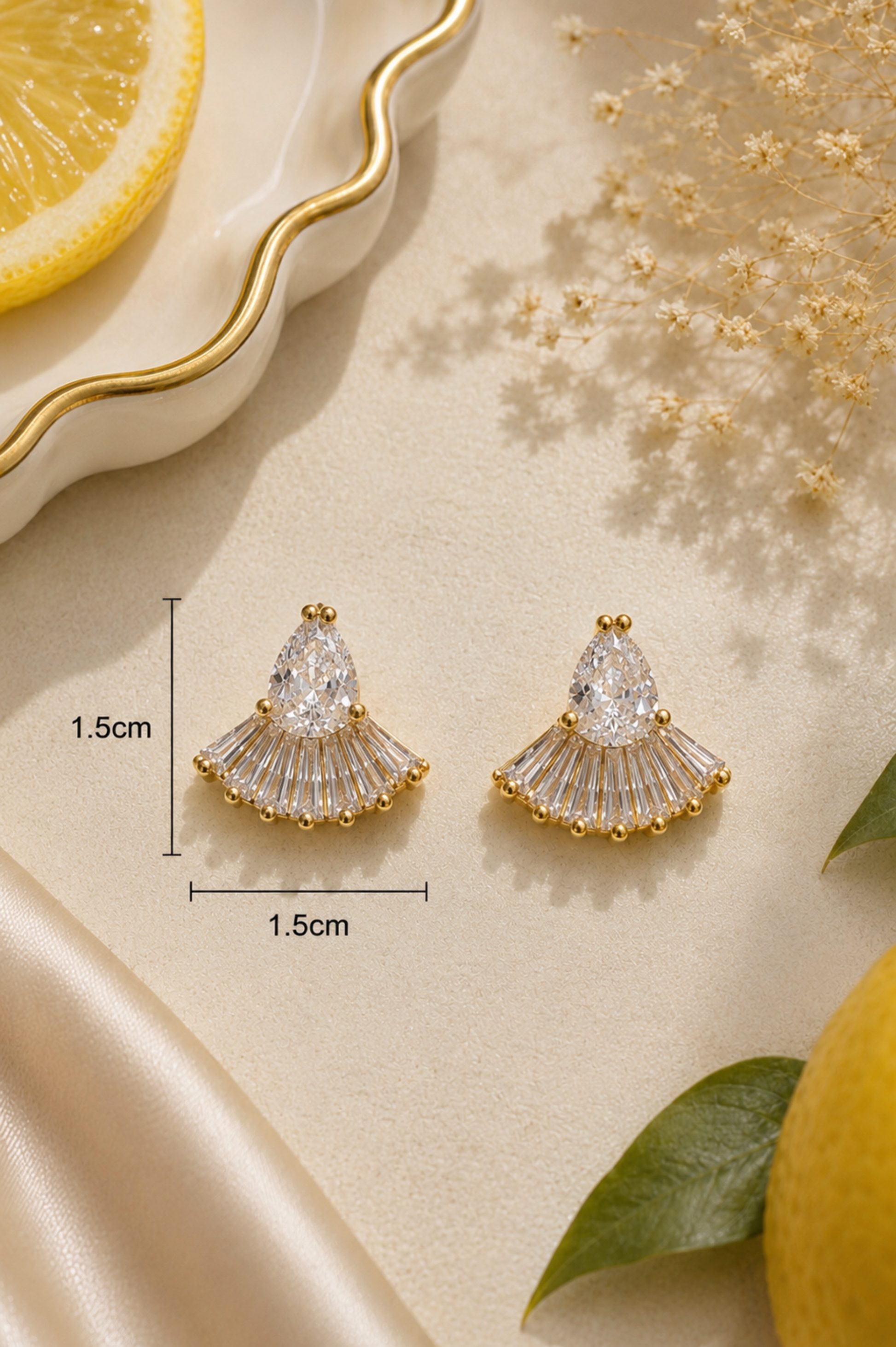 A pair of gold fan-shaped stud earrings with clear crystal-like drops, shown on a beige surface with decorative dried flowers, a lemon slice, green leaves, and measurement markers indicating 1.5cm width and height.