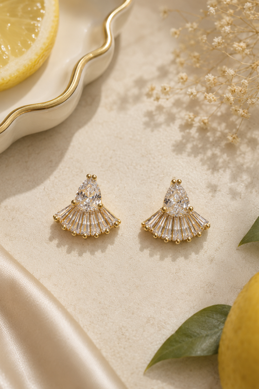 A pair of gold fan-shaped stud earrings with clear teardrop and baguette stones, styled on a beige surface with dried flowers, a lemon slice, green leaves, and a white dish with a gold rim.