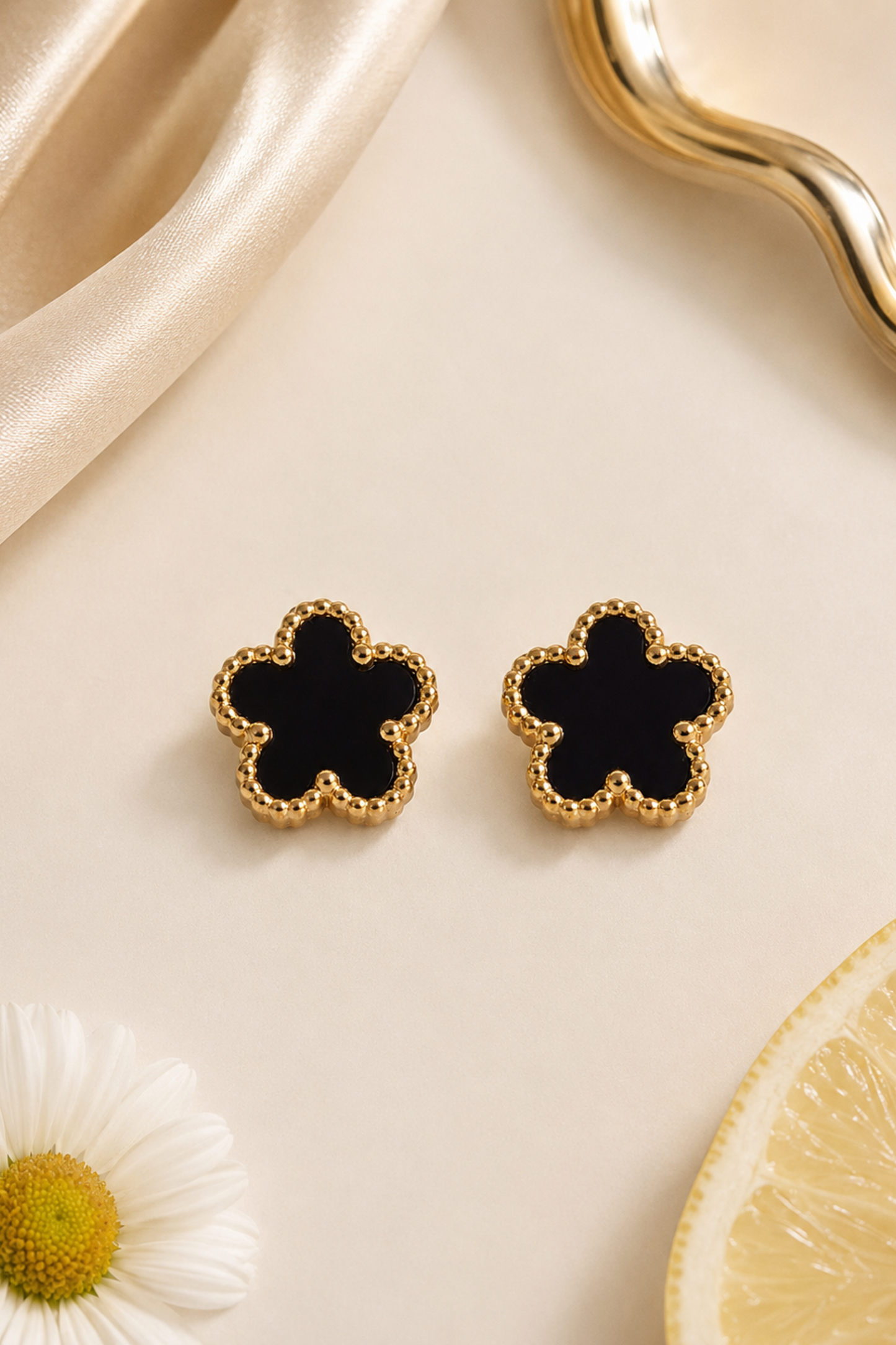 A pair of gold flower-shaped stud earrings with a black center, displayed on a beige surface with decorative satin, a daisy flower, and a lemon slice.