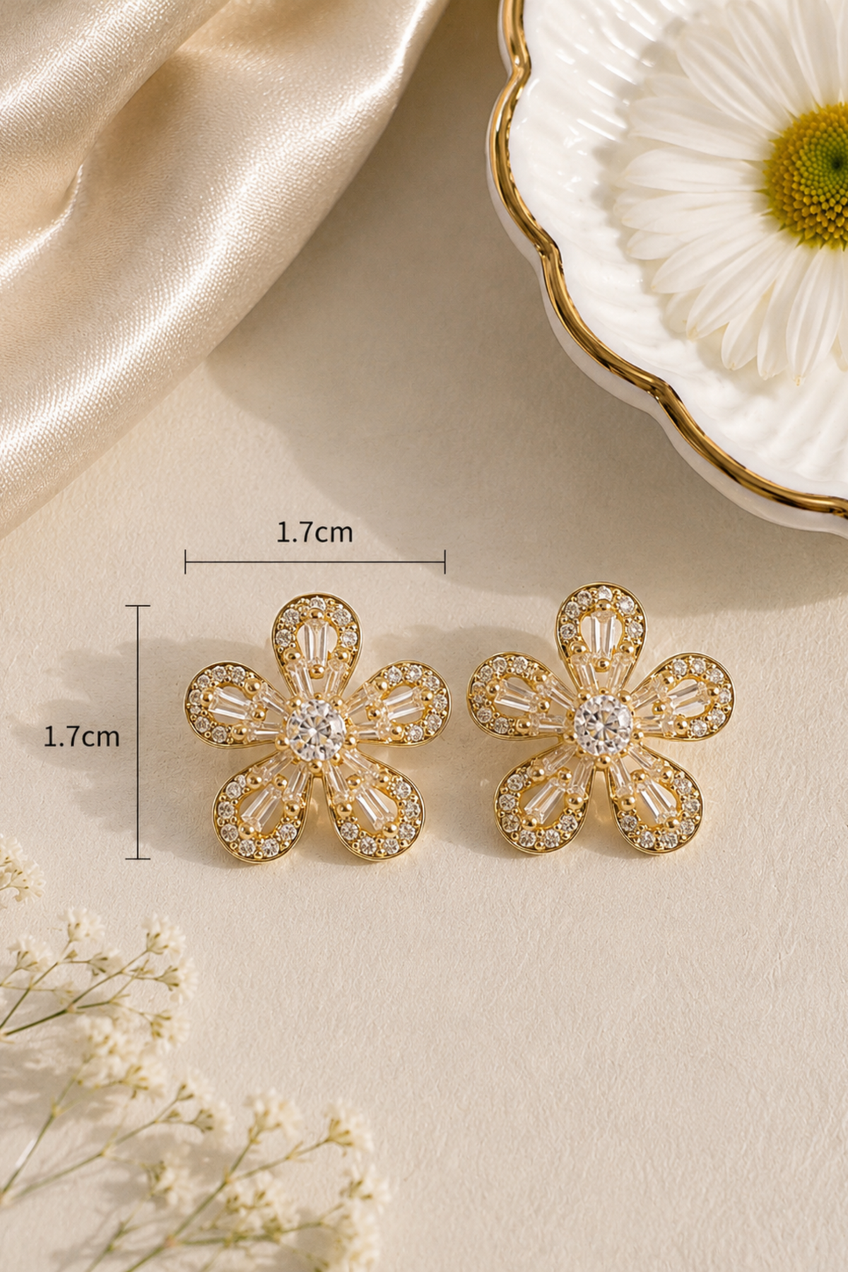 A pair of gold flower-shaped stud earrings with clear crystal details, displayed on a beige surface with dimension lines indicating a height and width of 1.7cm.