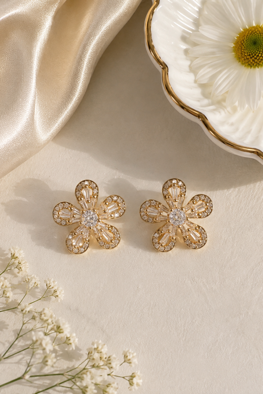 A pair of gold flower-shaped stud earrings with clear rhinestones, displayed on a textured cream surface with decorative satin fabric, a white daisy dish, and baby's breath flowers.