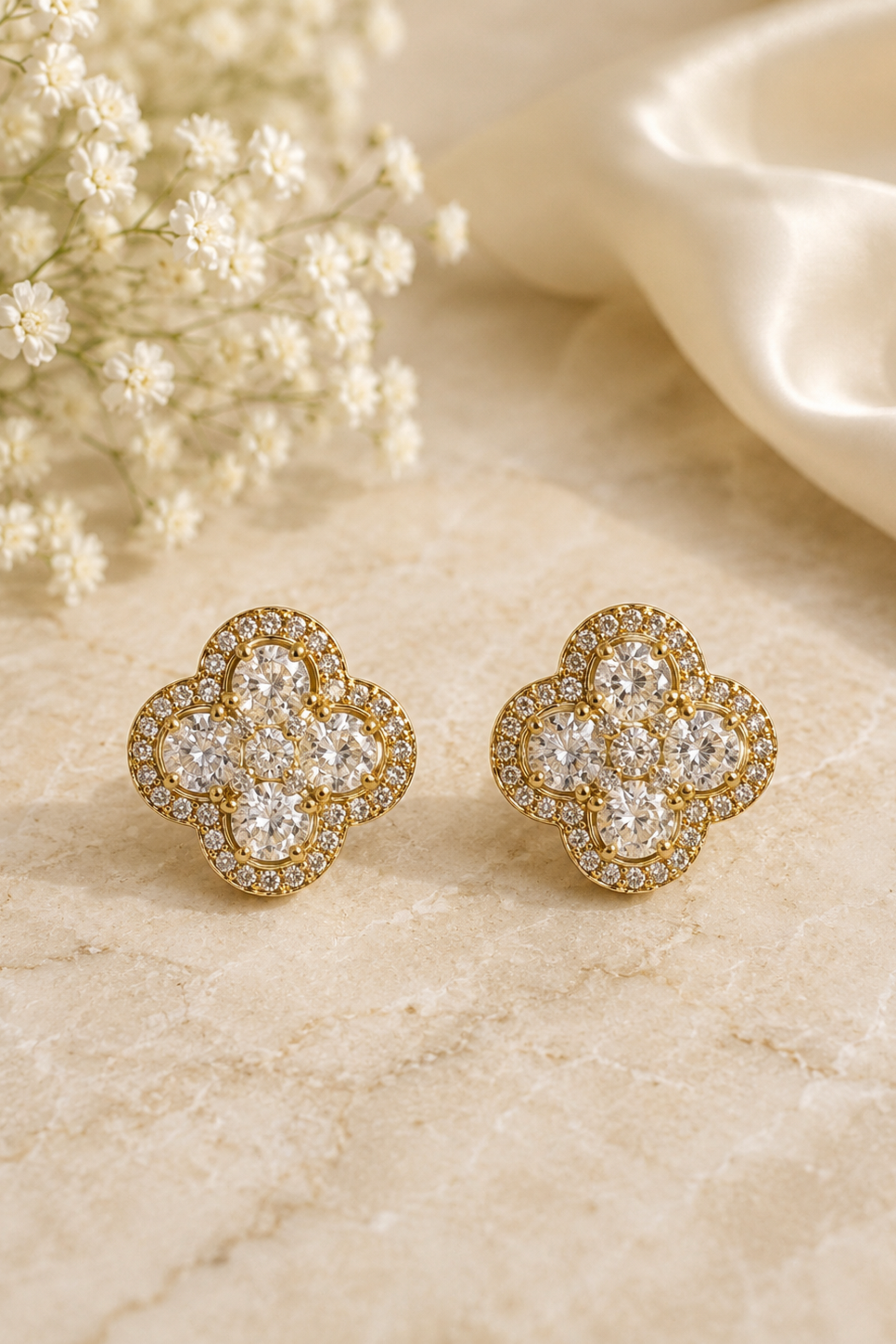 A pair of gold four-leaf clover stud earrings set with clear pavé gemstones, displayed on a beige marble surface next to white baby's breath flowers and a cream satin ribbon.