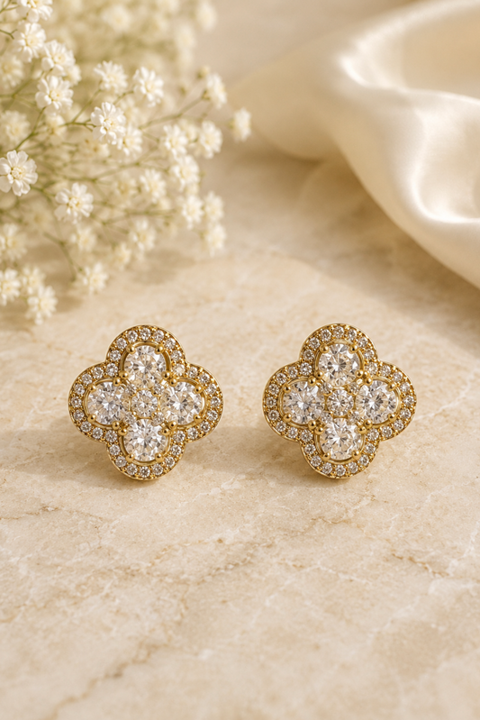 A pair of gold four-leaf clover stud earrings set with clear pavé gemstones, displayed on a beige marble surface next to white baby's breath flowers and a cream satin ribbon.