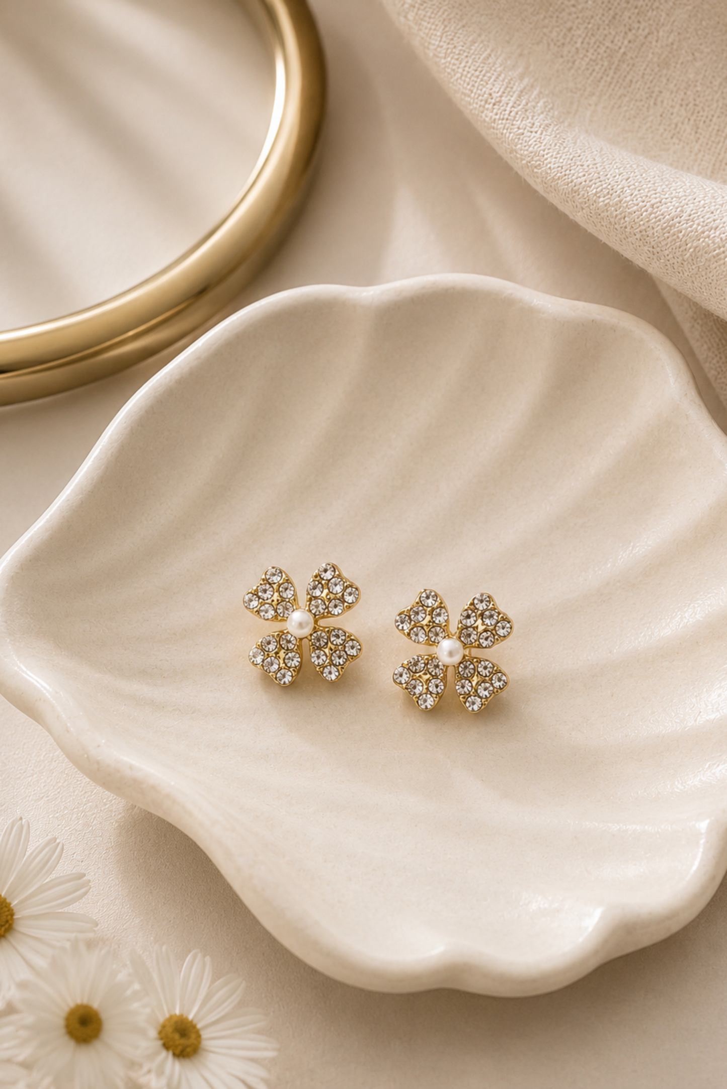 A pair of gold four-leaf clover stud earrings with clear rhinestones and a small white pearl in the center, displayed on a scalloped cream dish.