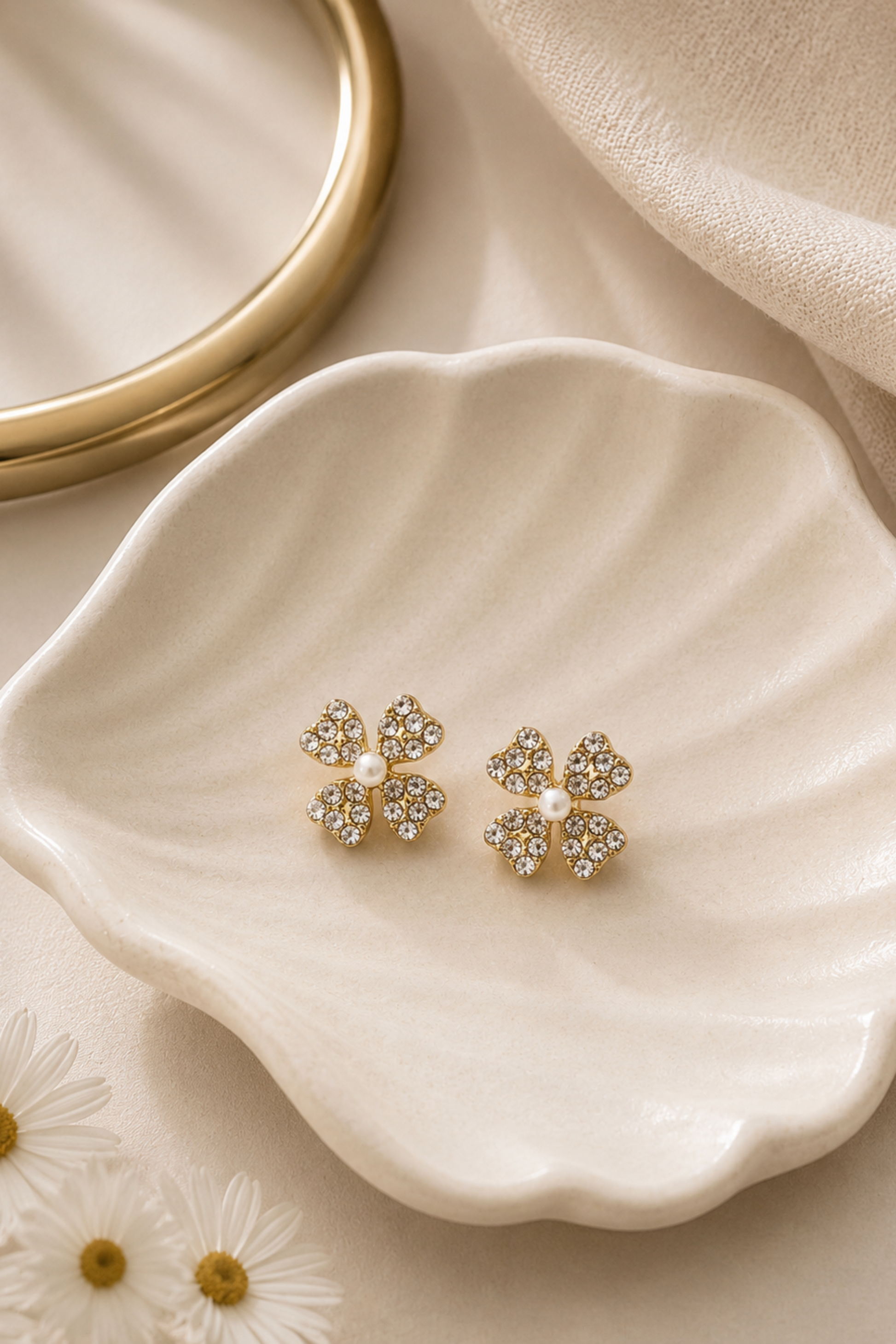 A pair of gold four-leaf clover stud earrings with clear rhinestones and a small white pearl in the center, displayed on a scalloped cream dish.