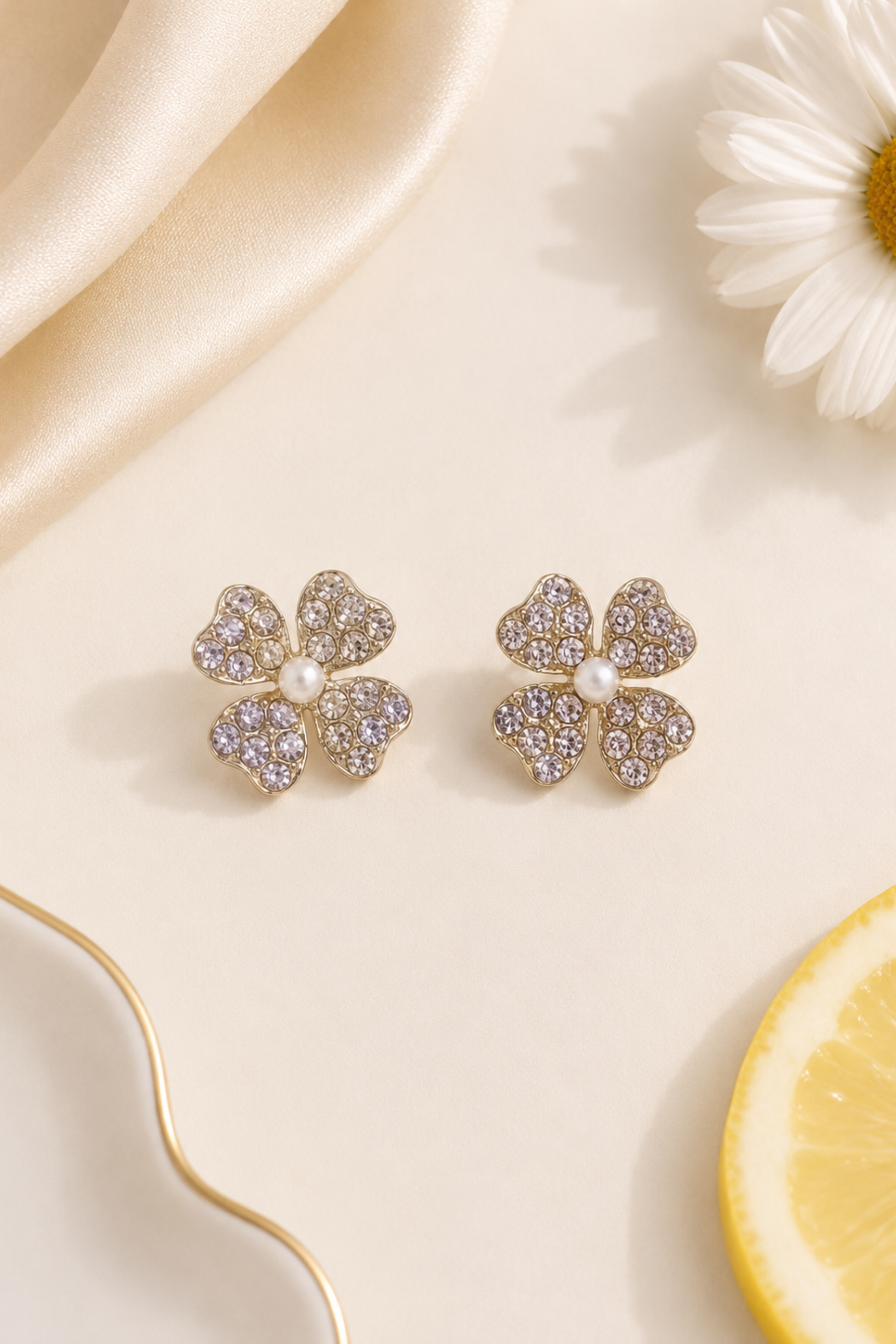 A pair of gold four-leaf clover stud earrings with pavé-set clear crystals and a small white pearl in the center, displayed on a cream surface with a daisy, satin fabric, and a lemon slice.