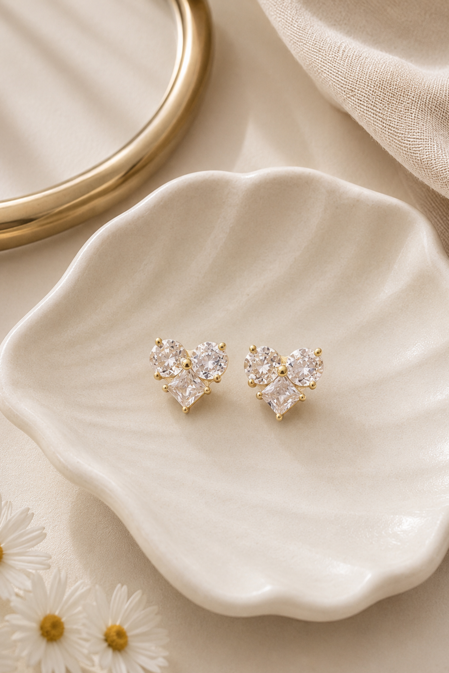 A pair of gold heart-shaped stud earrings with clear gemstones, displayed on a scalloped cream ceramic dish with daisies and a gold ring in the background.