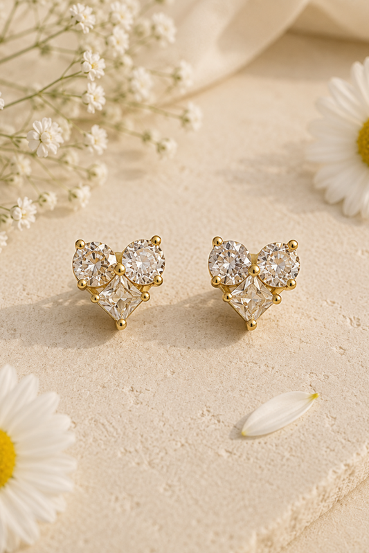 A pair of gold heart-shaped stud earrings with clear gemstones, styled on a textured beige surface with white daisies and soft cream fabric.