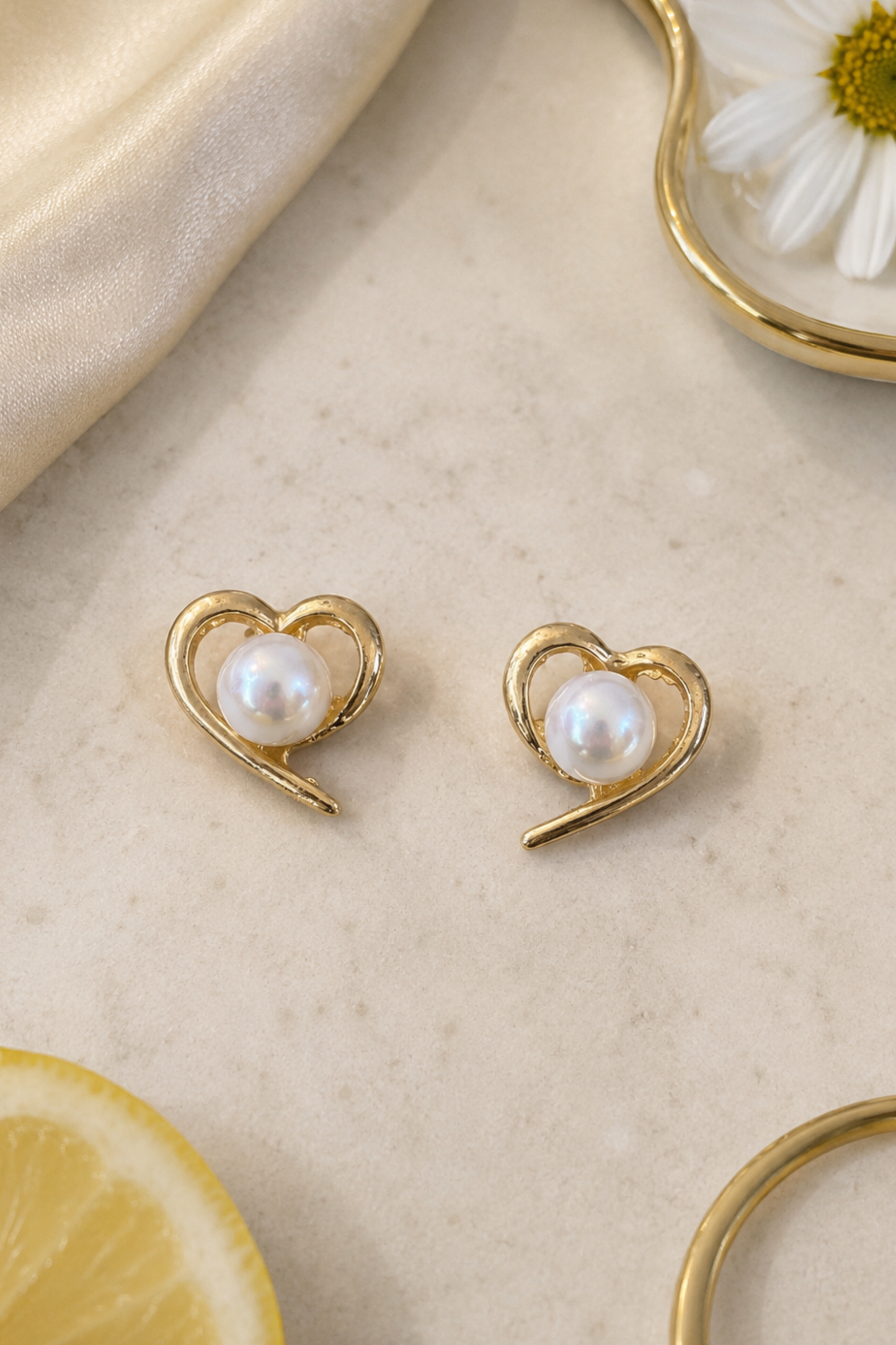 A pair of gold heart-shaped stud earrings with white freshwater pearls, displayed on a light beige marble surface with a cream satin cloth, a daisy flower, and a lemon slice.