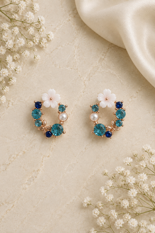 A pair of gold hoop-style earrings decorated with white flowers, blue and teal rhinestones, and small pearl-like beads, arranged on a textured cream surface with white baby's breath flowers and a draped cream fabric.