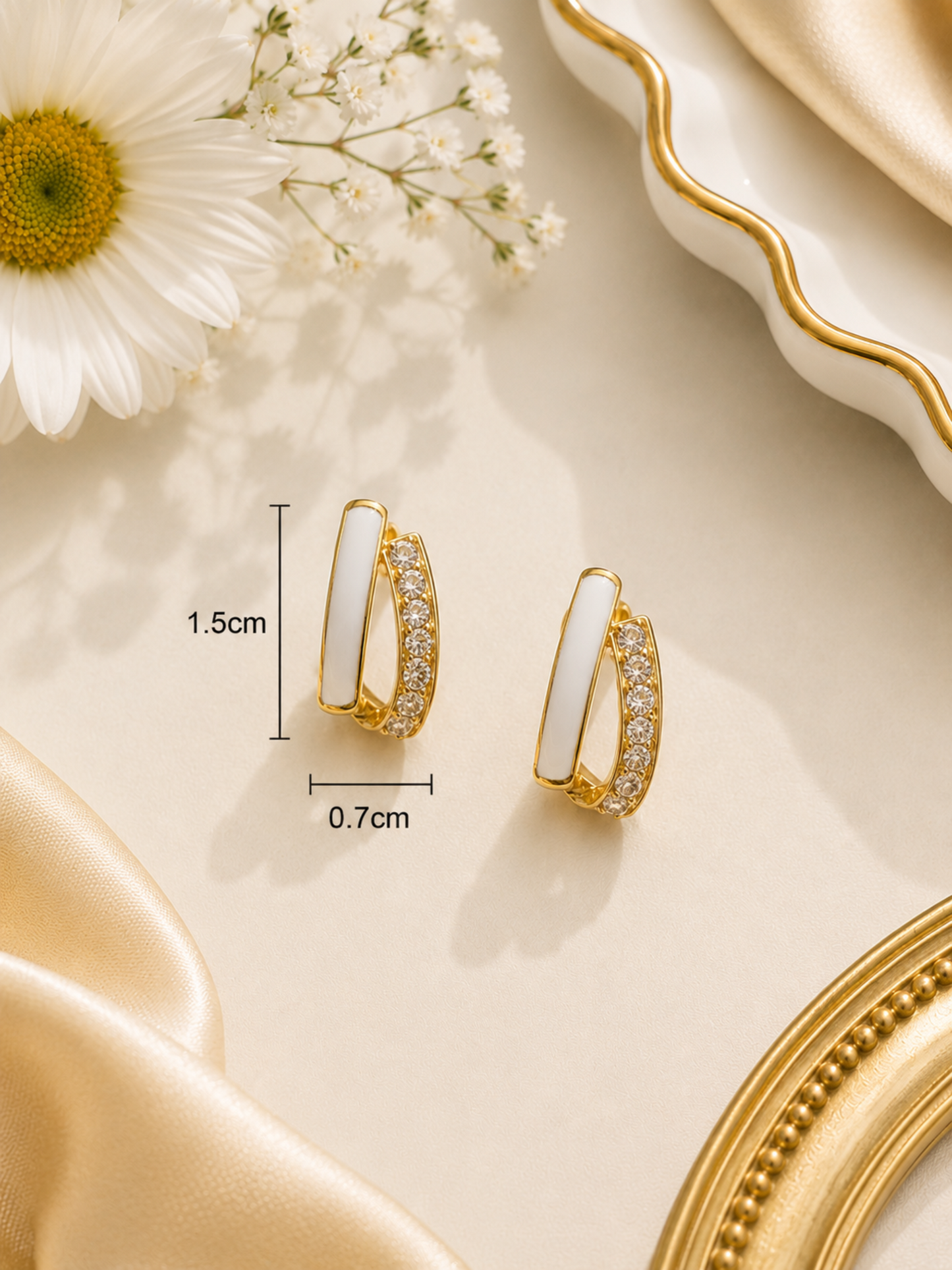 A pair of gold hoop-style earrings with white inlay and small clear stones, shown on a beige surface with decorative flowers, satin fabric, and a mirror edge. Measurement labels indicating 1.