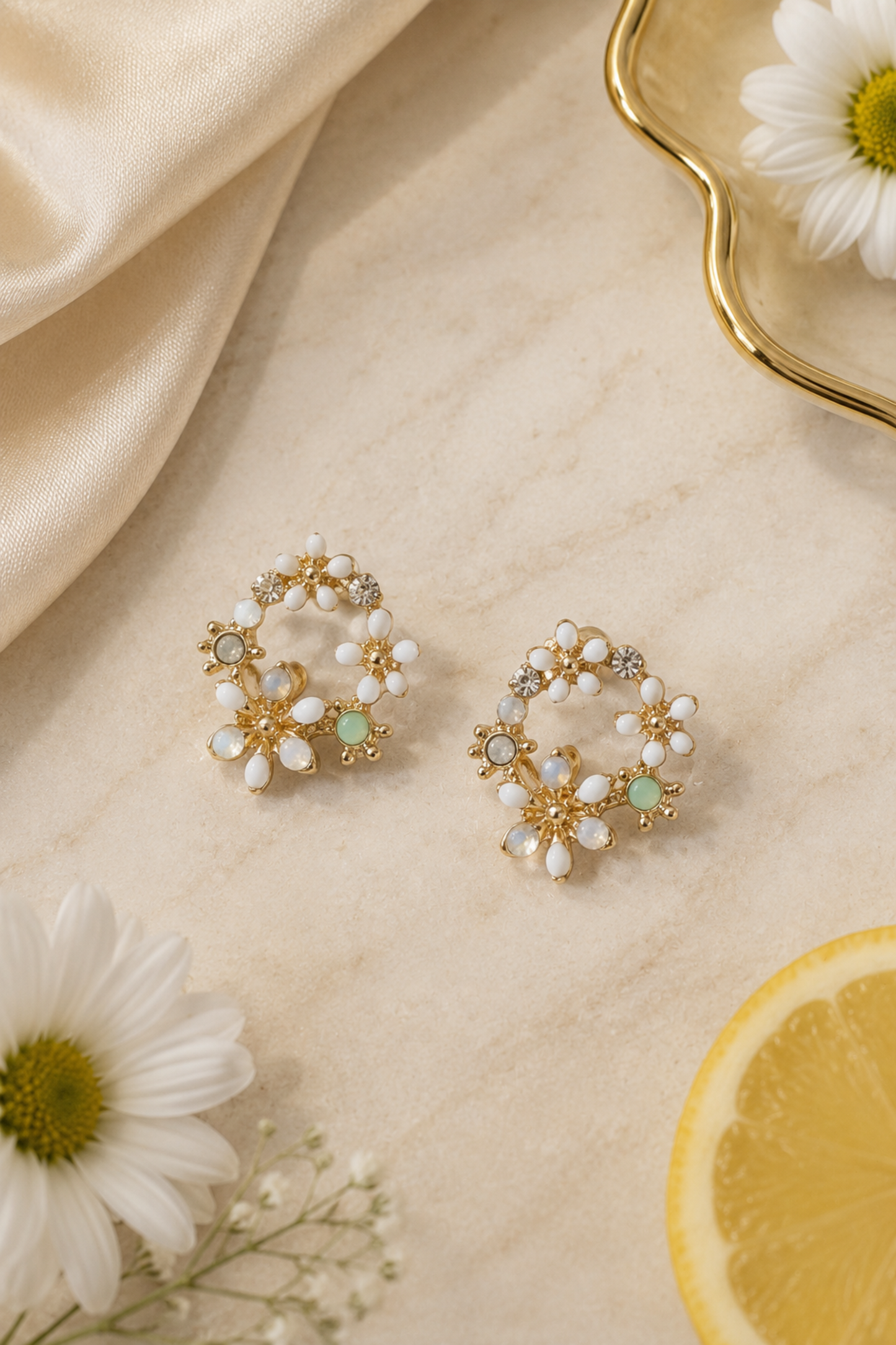 A pair of gold hoop earrings decorated with white faux pearls and small light green stones, styled on a beige marble surface with white daisies, a lemon slice, and draped satin fabric.
