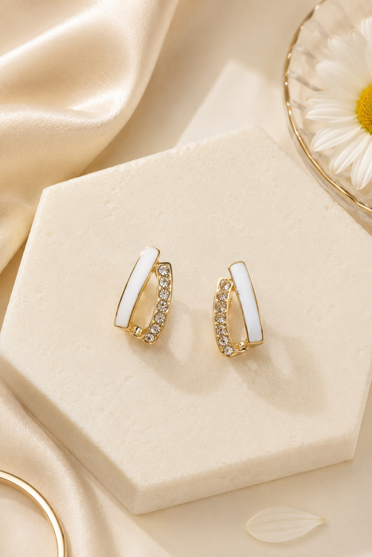 A pair of gold huggie hoop earrings with white inlay and clear crystal accents, displayed on a cream-colored hexagonal stand with a satin fabric and a daisy flower in the background.