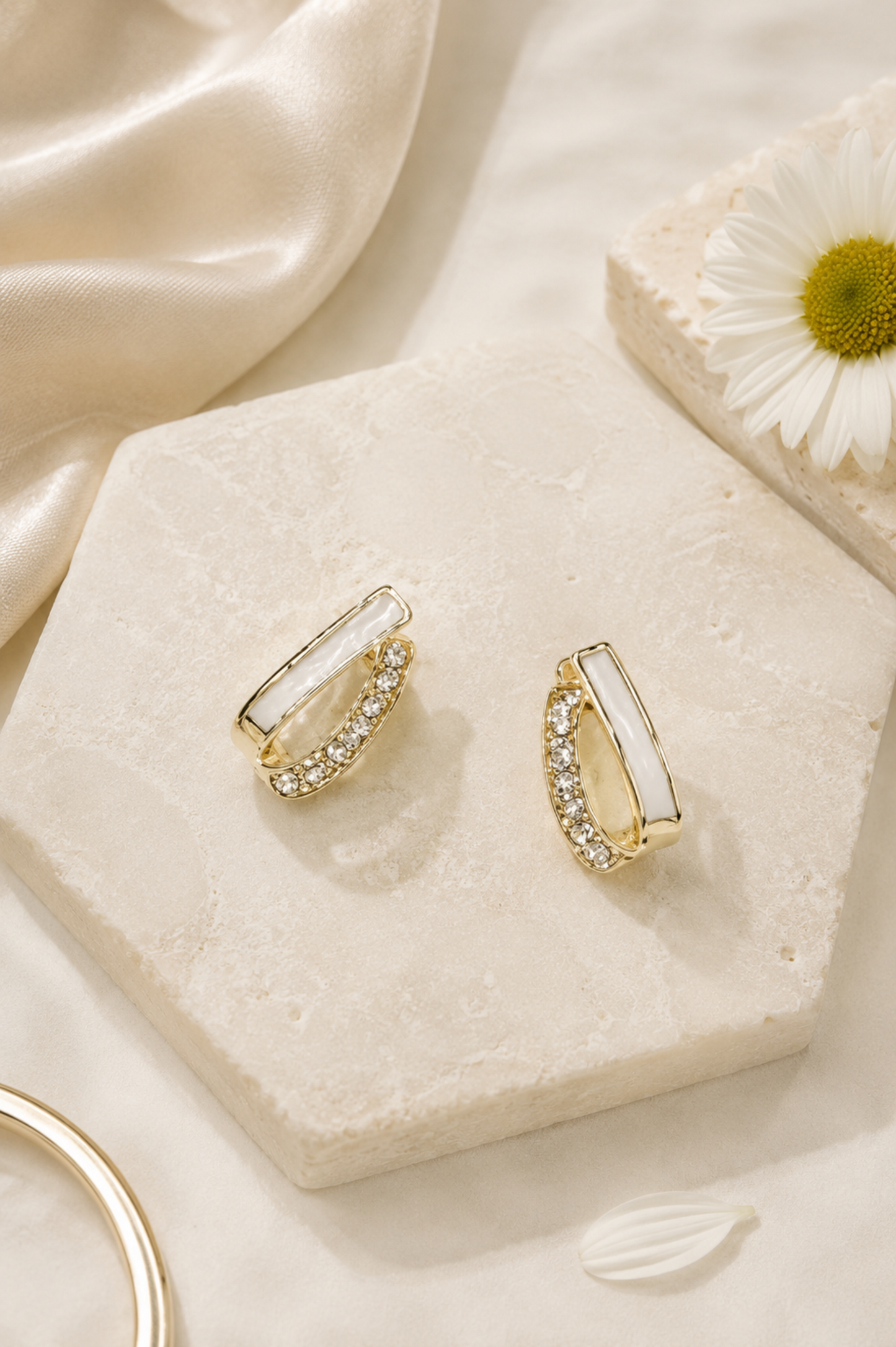 A pair of gold huggie hoop earrings with white inlay and clear crystal accents, displayed on a textured cream stone hexagon with a white satin fabric and a daisy flower in the background.