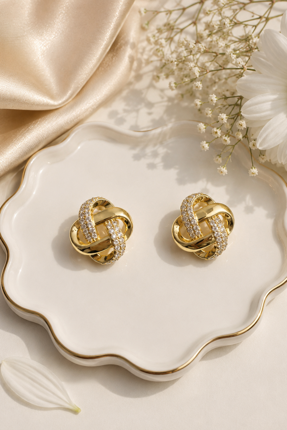A pair of gold knot-shaped stud earrings with pavé-set clear stones, displayed on a scalloped white ceramic dish with gold trim, surrounded by champagne satin fabric, baby's breath, and a white daisy.