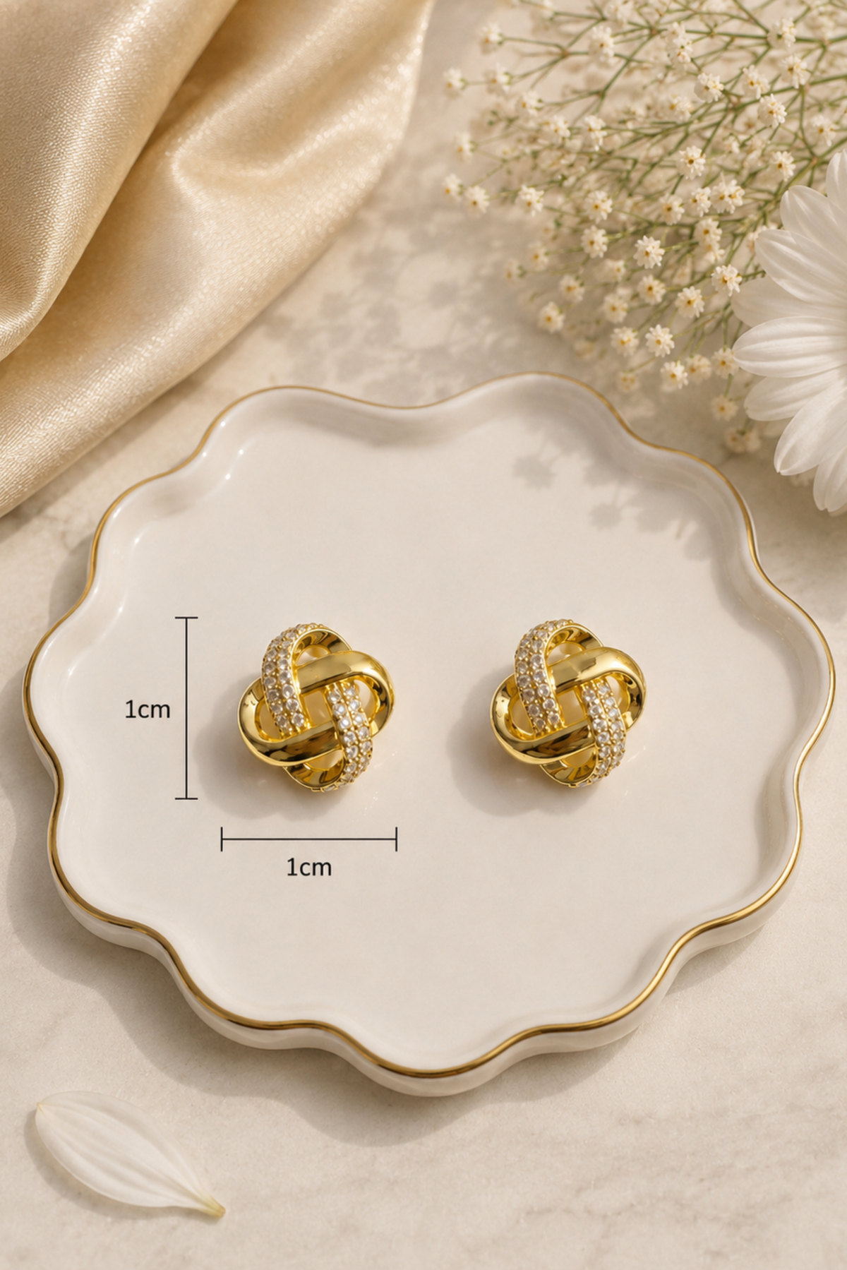 A pair of gold knot-shaped stud earrings with small clear stones, displayed on a scalloped white ceramic dish with size markers indicating 1cm by 1cm. The dish is styled with gold satin fabric, baby's breath flowers, and a white daisy on a light marble surface.