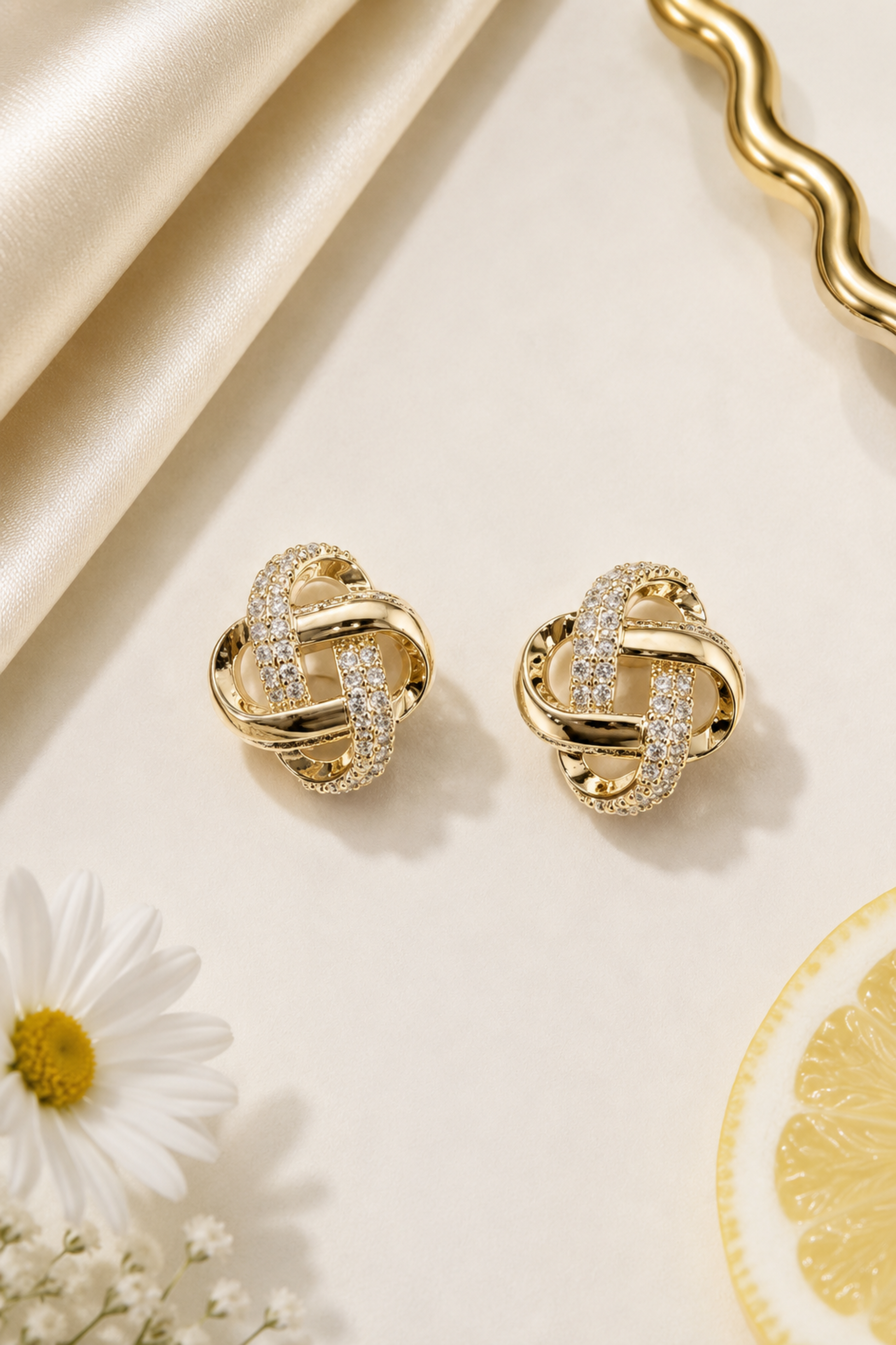 A pair of gold knot stud earrings with pavé-set clear stones, displayed on a cream-colored surface with a cream satin ribbon, a white daisy flower, and a lemon slice.