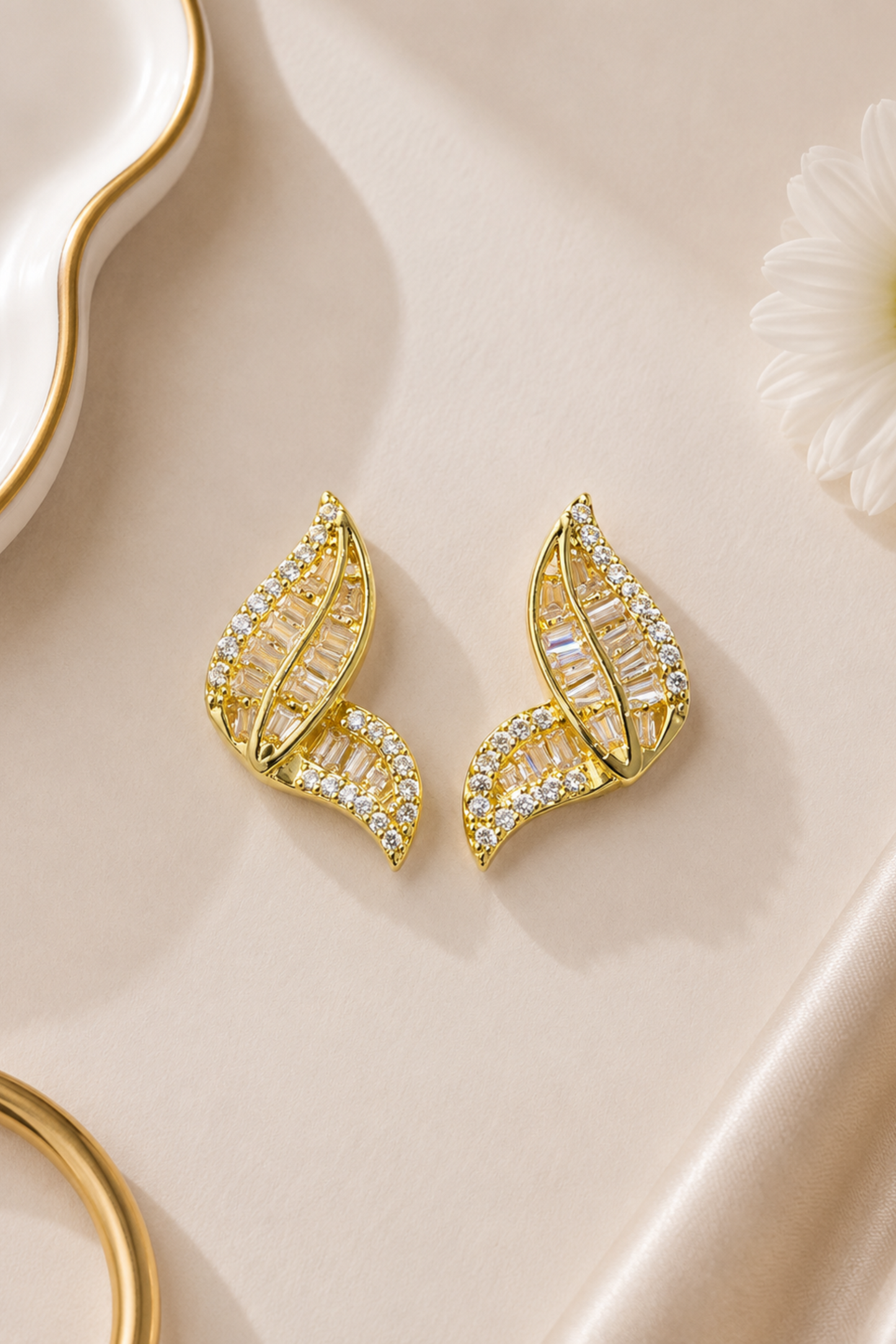 A pair of gold leaf-shaped stud earrings adorned with clear baguette and round crystals, displayed on a beige satin fabric background with decorative gold and white props.