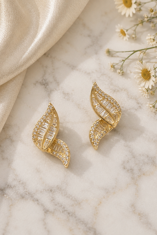 A pair of gold leaf-shaped stud earrings adorned with clear baguette and round crystals, displayed on a white marble surface next to a draped champagne satin cloth and small white daisies.