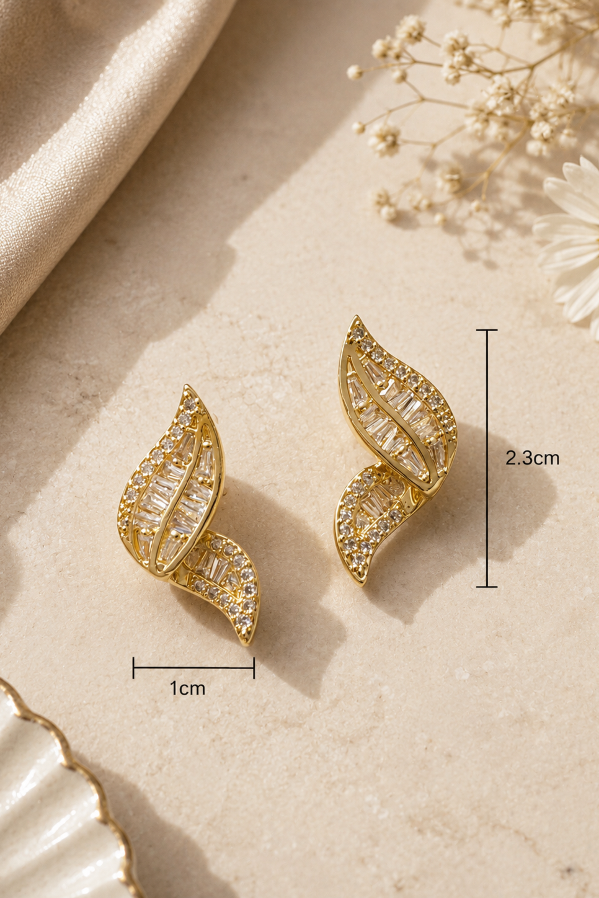 A pair of gold leaf-shaped stud earrings with clear crystal detailing, displayed on a beige textured surface with decorative dried flowers and a draped fabric. Black measurement lines and text indicate dimensions of 1cm width, 2.