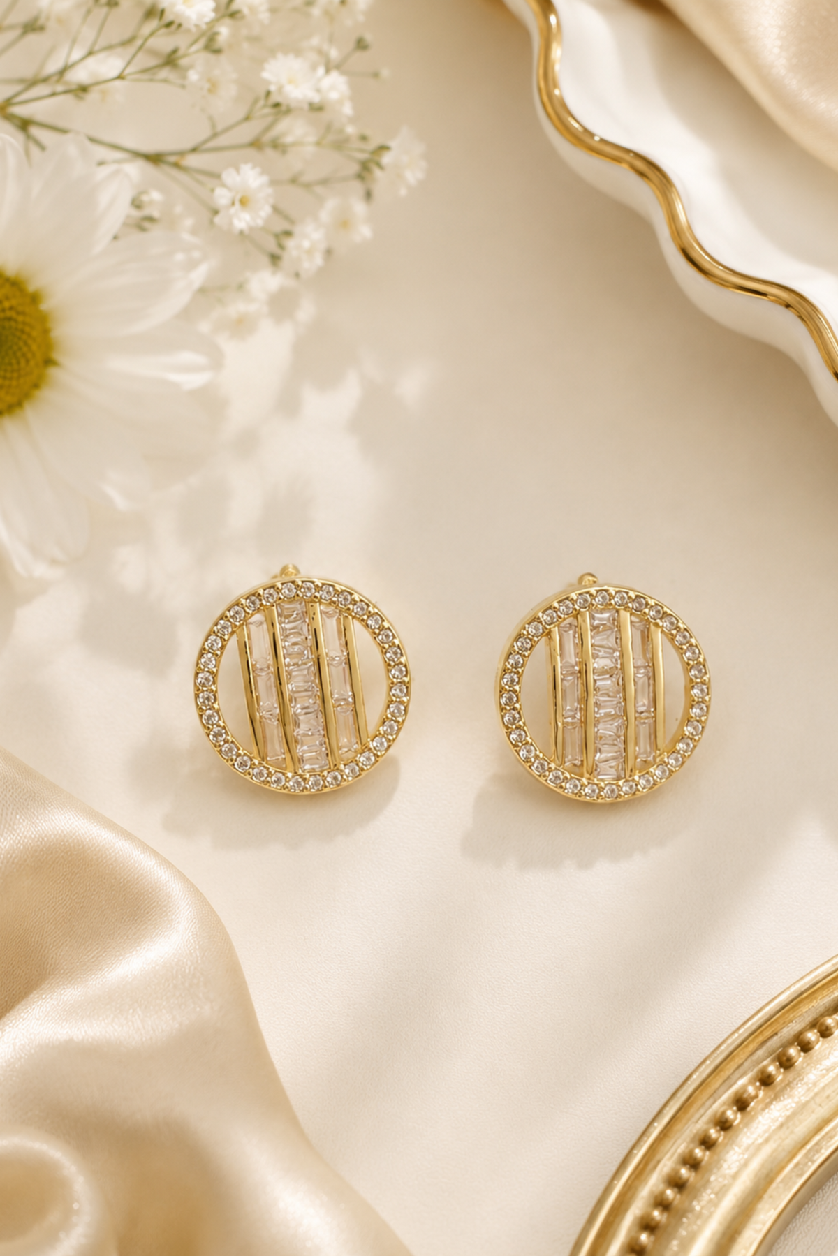 A pair of gold oval stud earrings with vertical bar details and clear stones, displayed on a cream satin surface with a daisy flower and decorative gold-toned props.