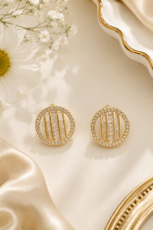 A pair of gold oval stud earrings with vertical bar details and clear stones, displayed on a cream satin surface with a daisy flower and decorative gold-toned props.