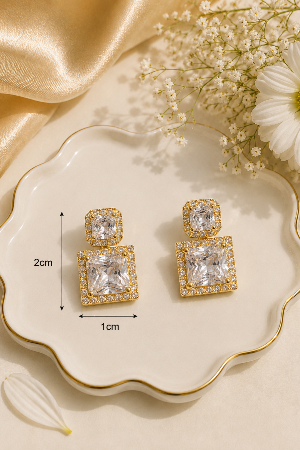 A pair of gold square drop earrings with clear gemstones, displayed on a scalloped cream dish with measurement labels indicating 2cm height and 1cm width. The scene is styled with gold satin fabric, baby's breath flowers, and a white daisy.