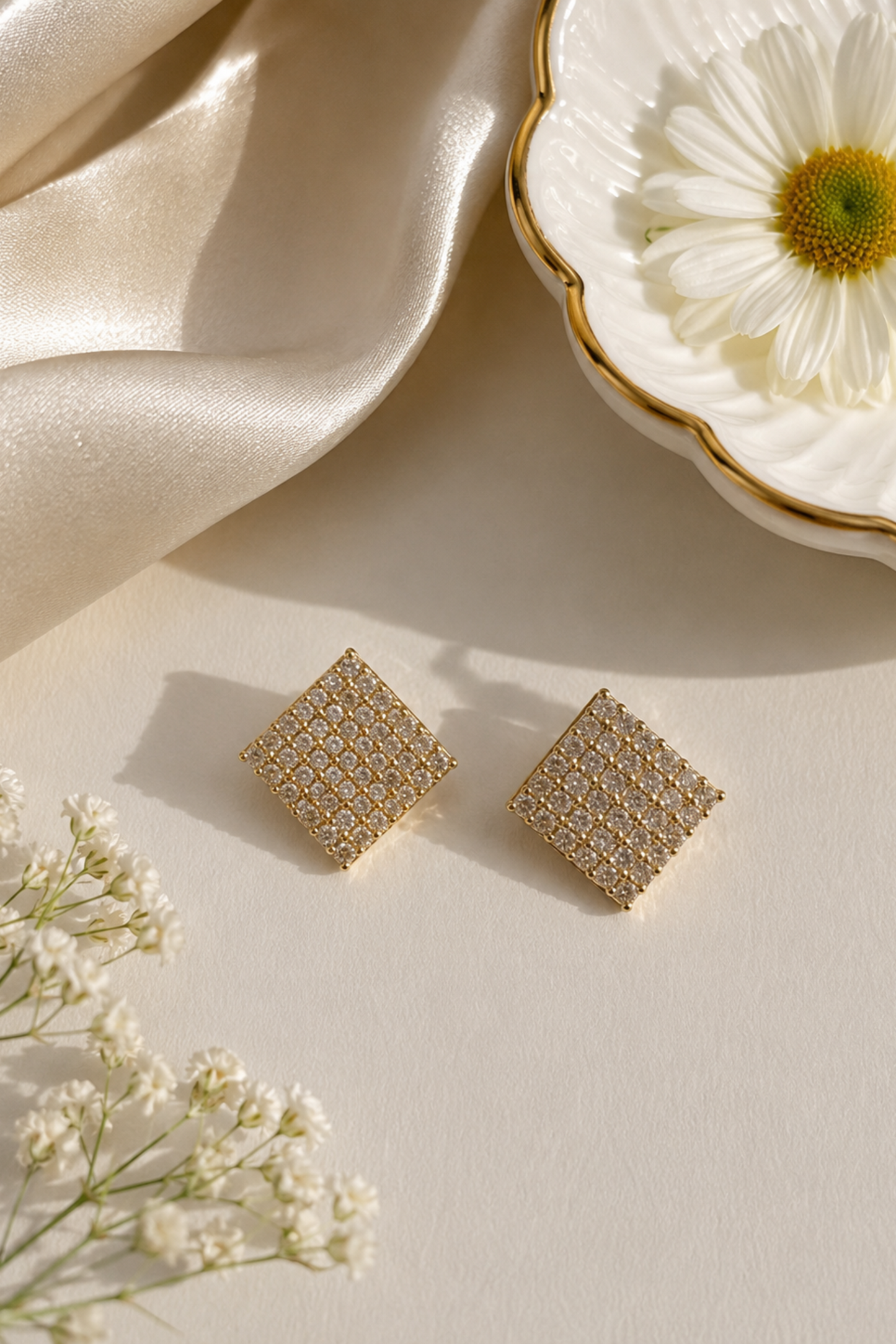 A pair of gold square stud earrings with a textured, openwork geometric pattern, styled on a cream surface with a white daisy, baby's breath flowers, and draped champagne-colored satin fabric.