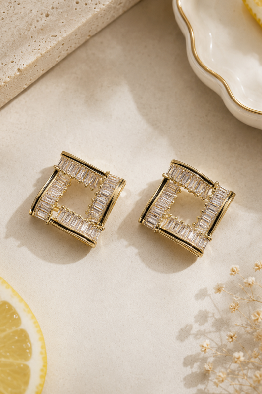 A pair of gold square stud earrings with baguette-cut clear stones, styled on a light beige surface with lemon slices and dried flowers.
