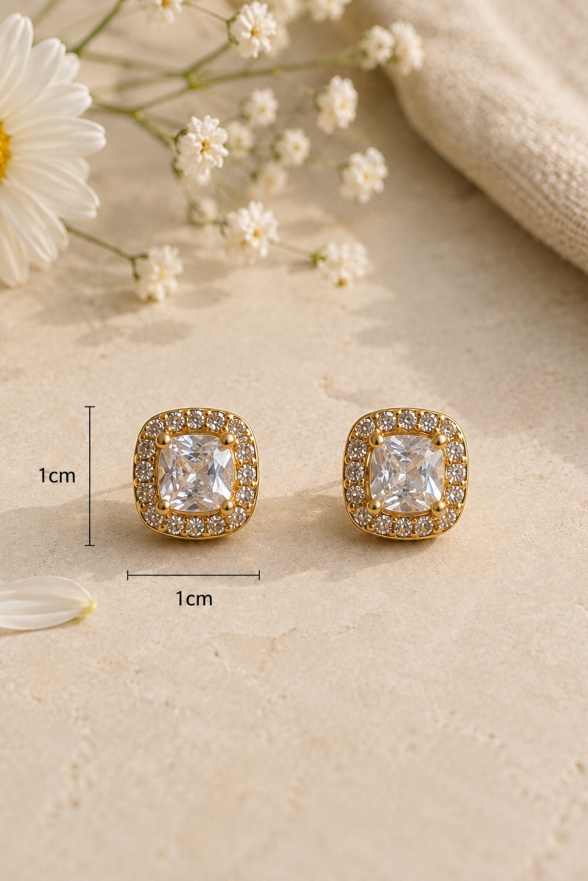 A pair of gold square stud earrings with clear cushion-cut center stones and a halo of small clear stones, shown on a beige surface with decorative flowers and a fabric corner. 1cm size markers are overlaid near the earrings.
