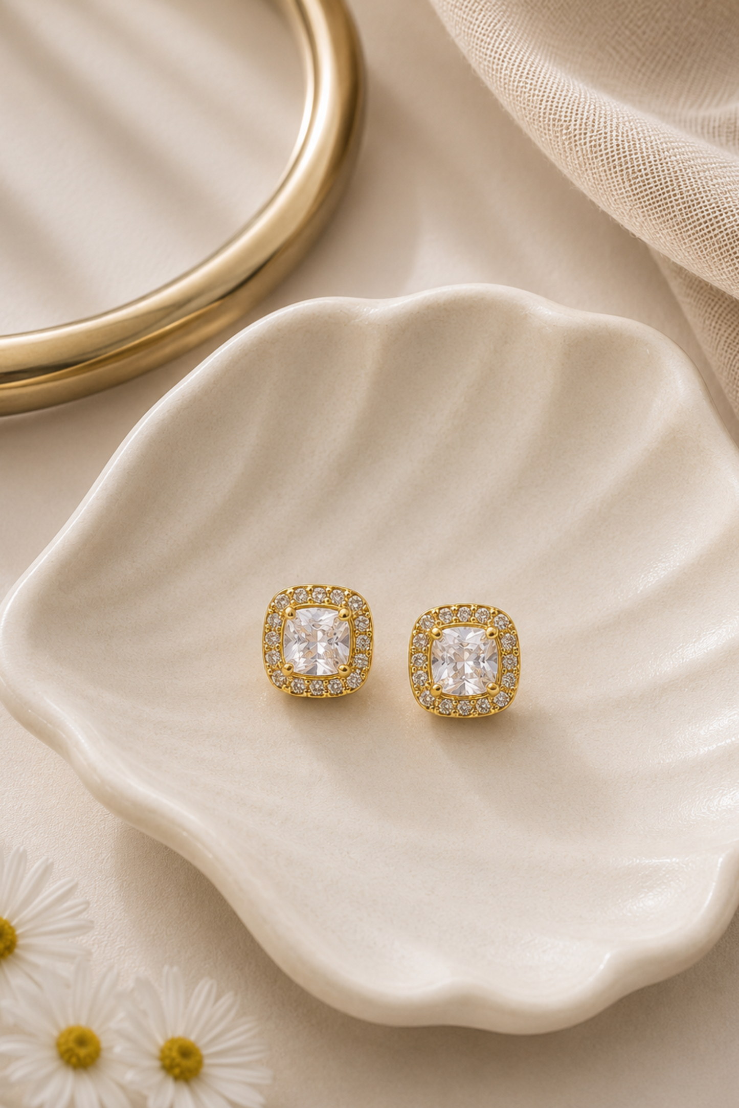 A pair of gold square stud earrings with clear cushion-cut center stones and small clear stones around the edges, displayed on a cream scalloped dish with a gold ring and beige fabric in the background.