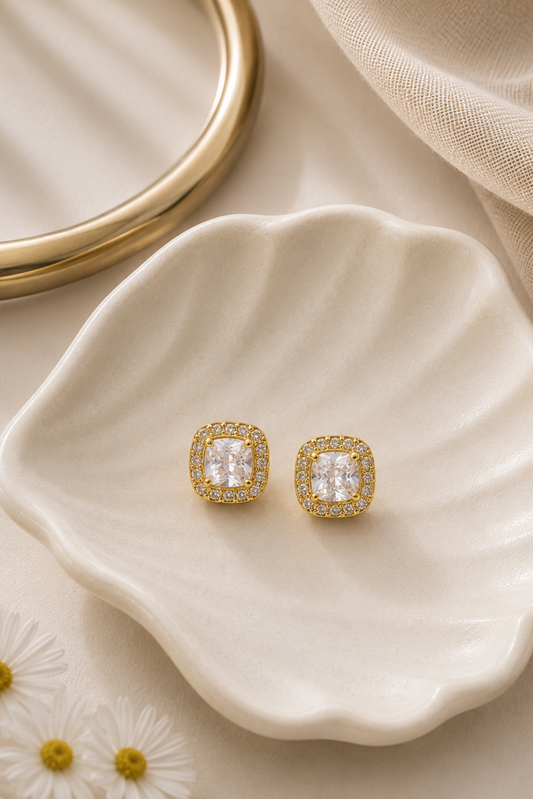 A pair of gold square stud earrings with clear cushion-cut center stones and small clear stones around the edges, displayed on a cream scalloped dish with a gold ring and beige fabric in the background.