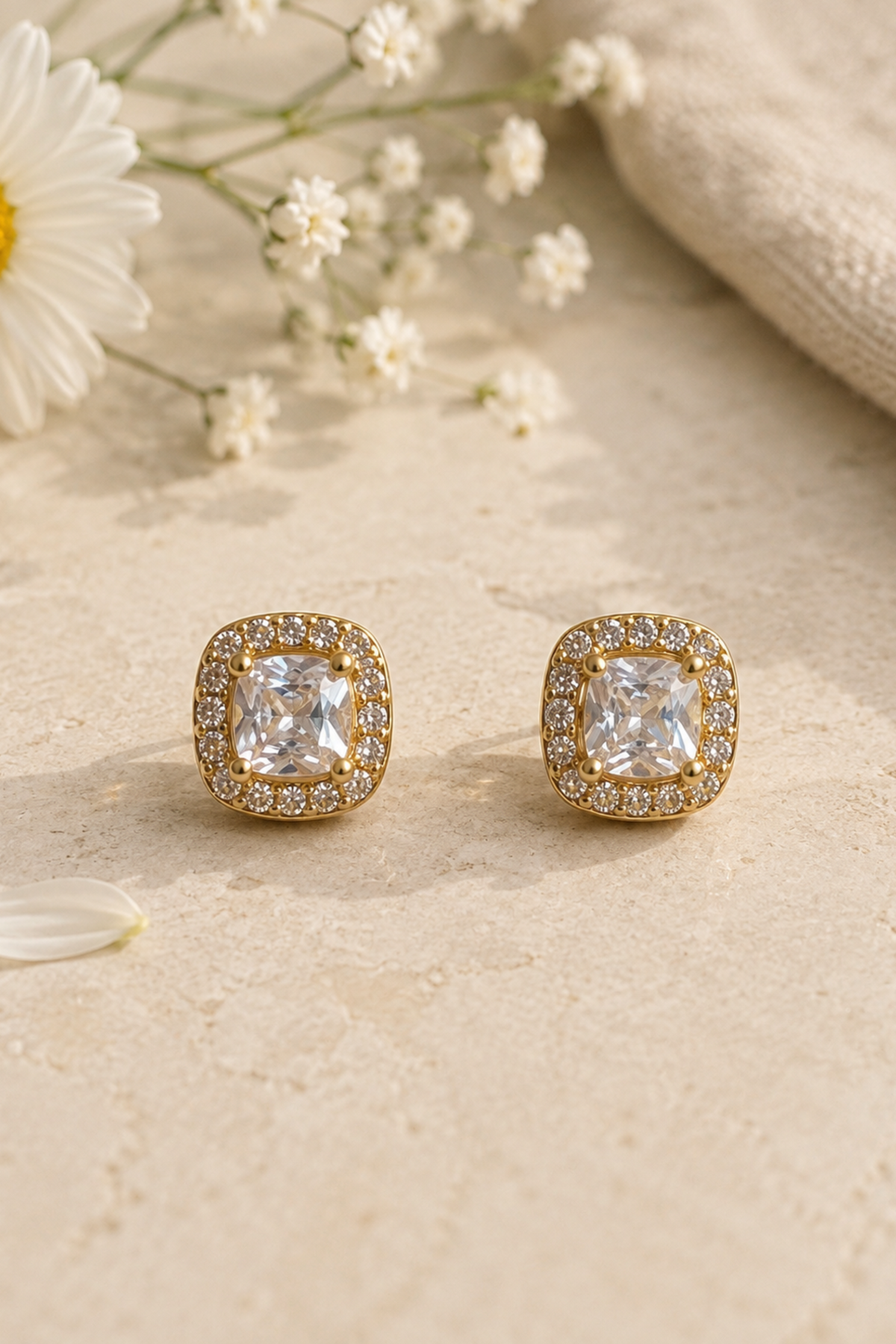 A pair of gold square stud earrings with cushion-cut clear gemstones and a halo of small clear stones, displayed on a textured beige surface with a daisy and baby's breath flowers.