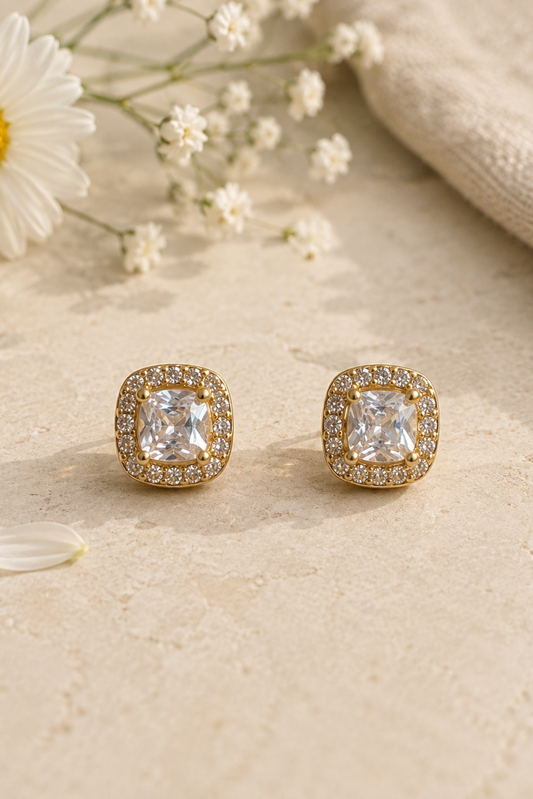 A pair of gold square stud earrings with cushion-cut clear gemstones and a halo of small clear stones, displayed on a textured beige surface with a daisy and baby's breath flowers.