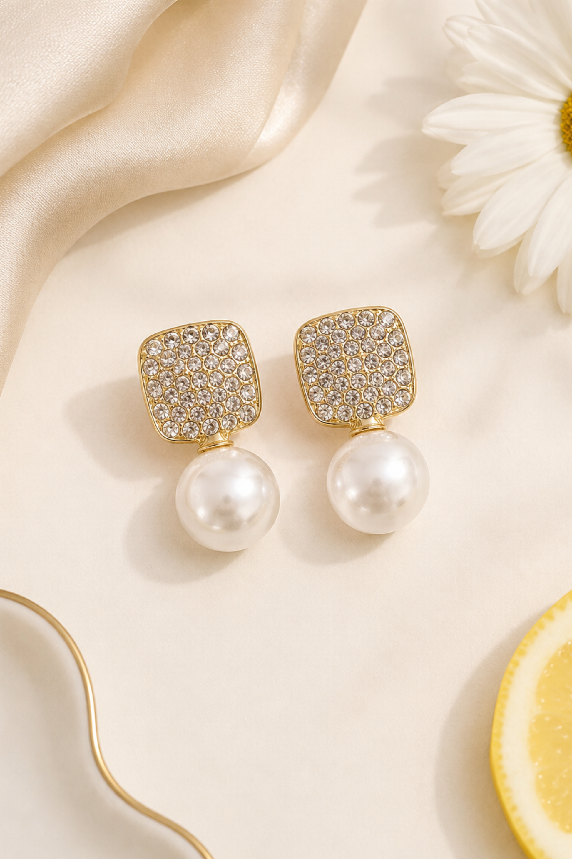 A pair of gold square stud earrings with pavé-set clear stones and dangling white pearls, styled on a cream surface with satin fabric, a daisy flower, and a lemon slice.