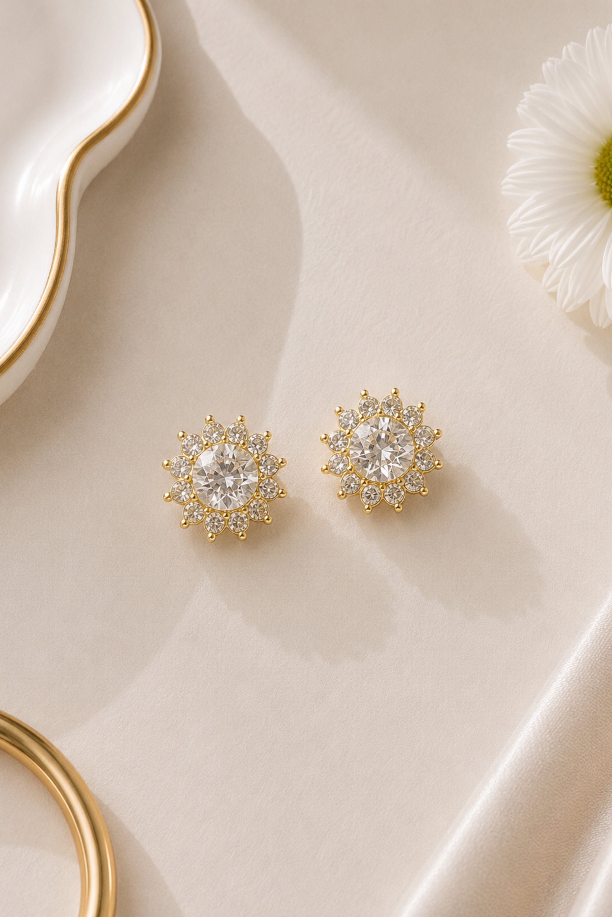 A pair of gold starburst stud earrings with clear gemstones, displayed on a beige satin fabric with decorative props.
