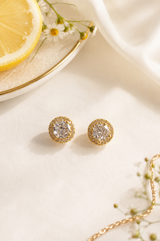 A pair of gold stud earrings with clear pavé-set round stones, displayed on a light cream surface with a lemon slice, white daisies, and a gold chain as styled props.