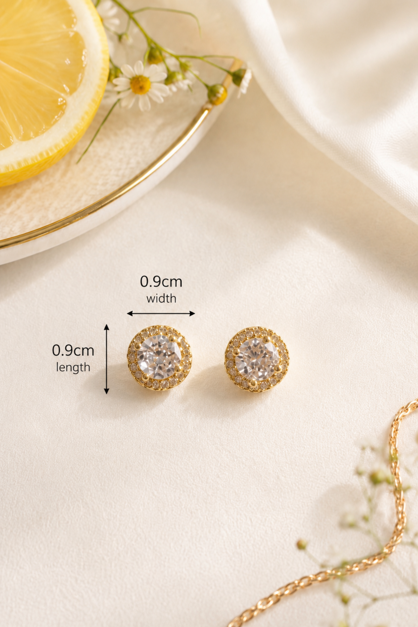 A pair of gold stud earrings with clear round gemstones, shown on a light cream fabric surface with measurement arrows and text indicating 0.9cm width and 0.