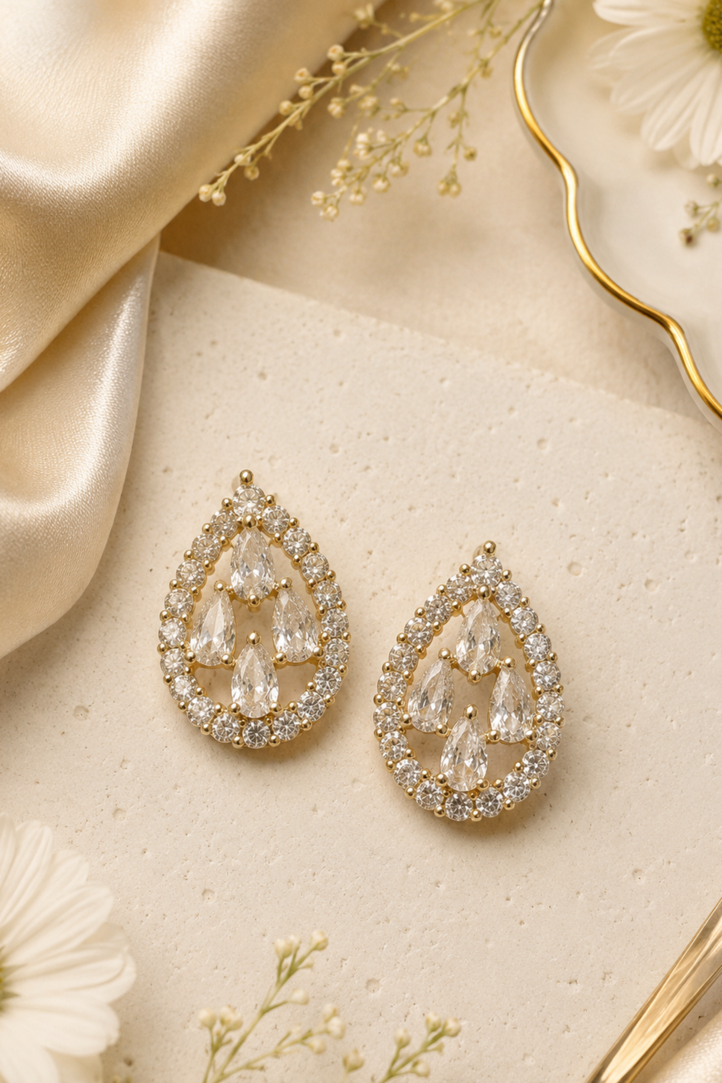 A pair of gold teardrop-shaped drop earrings with clear teardrop and round crystals, arranged on a textured cream surface with gold ribbon, white daisies, and baby's breath flowers.