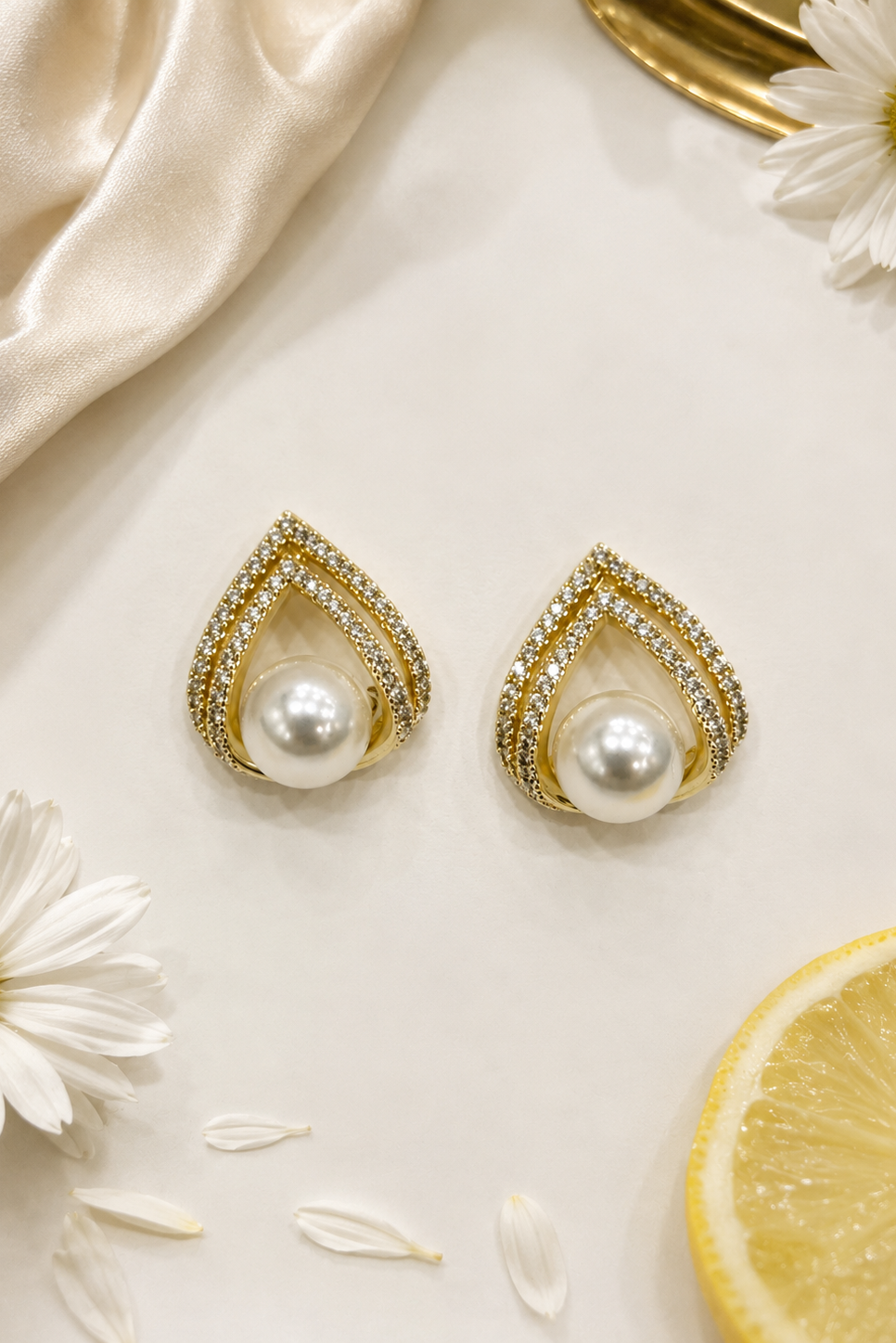 A pair of gold teardrop-shaped earrings with a central white pearl and a border of small clear stones, styled on a cream surface with white daisies, a lemon slice, and draped satin fabric.