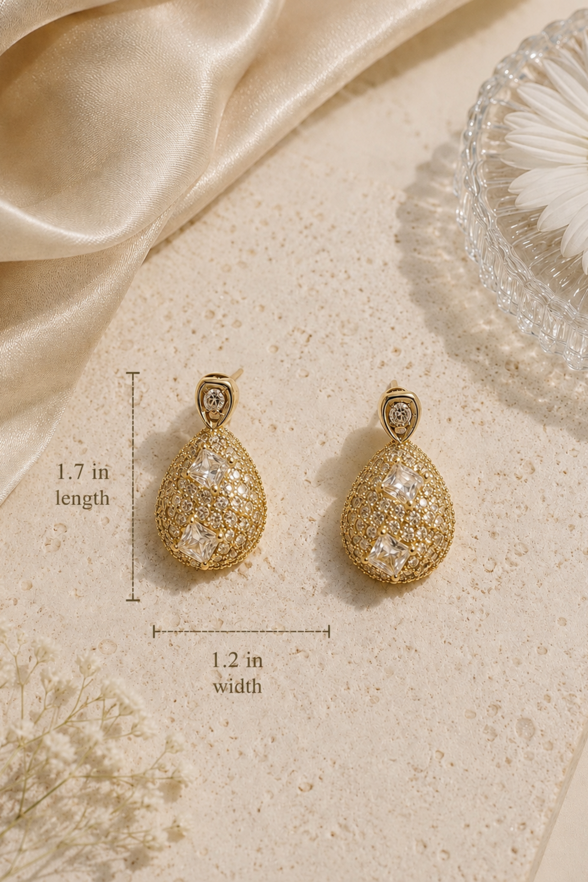 A pair of gold teardrop-shaped earrings with sparkling clear stones, displayed on a textured beige surface with decorative satin fabric, a daisy flower, and dimension lines showing 1.7 in length and 1.