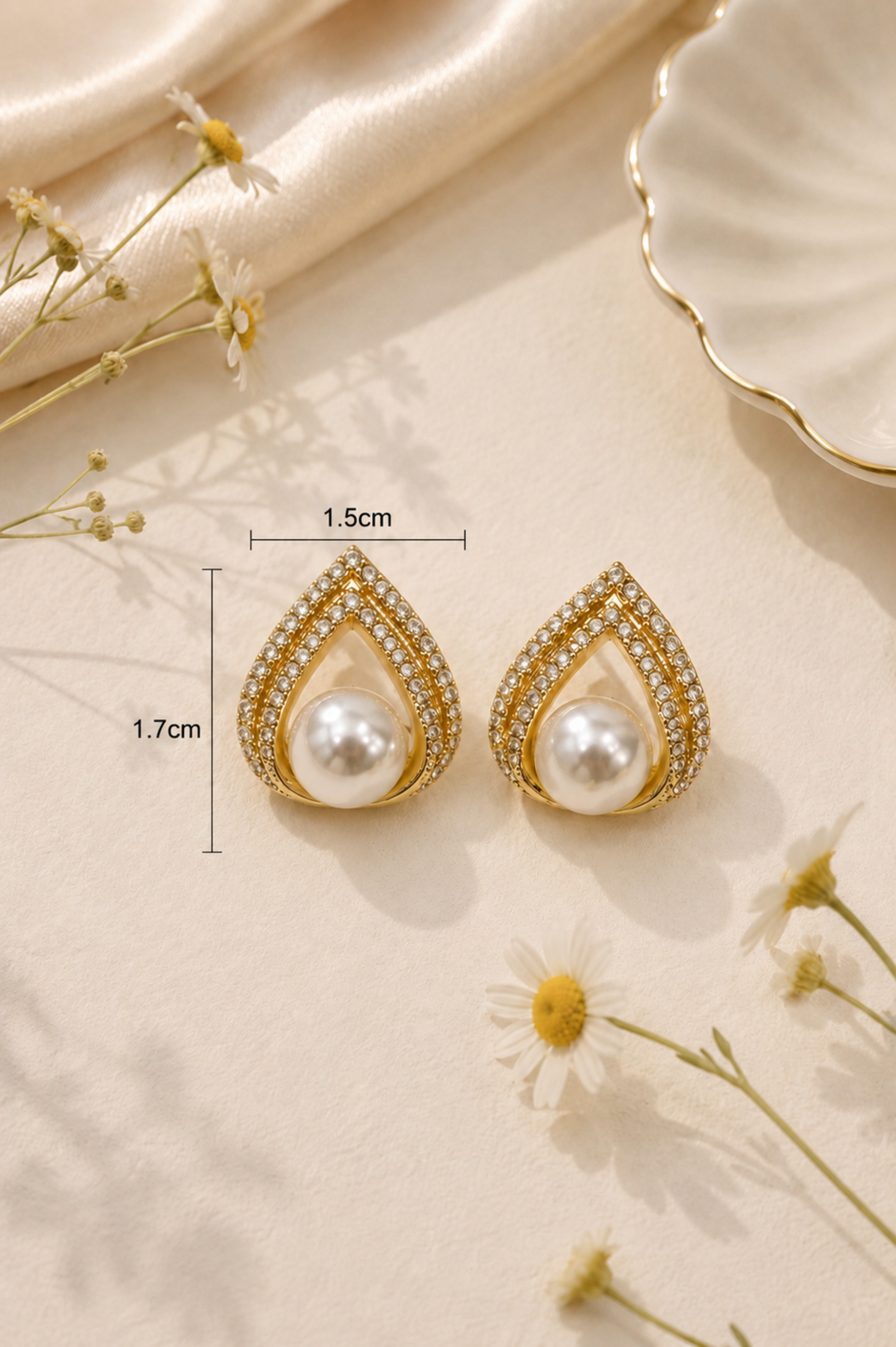 A pair of gold teardrop-shaped earrings with white pearls and small clear stones, shown on a beige surface with dried flowers and fabric. Measurement lines indicate 1.