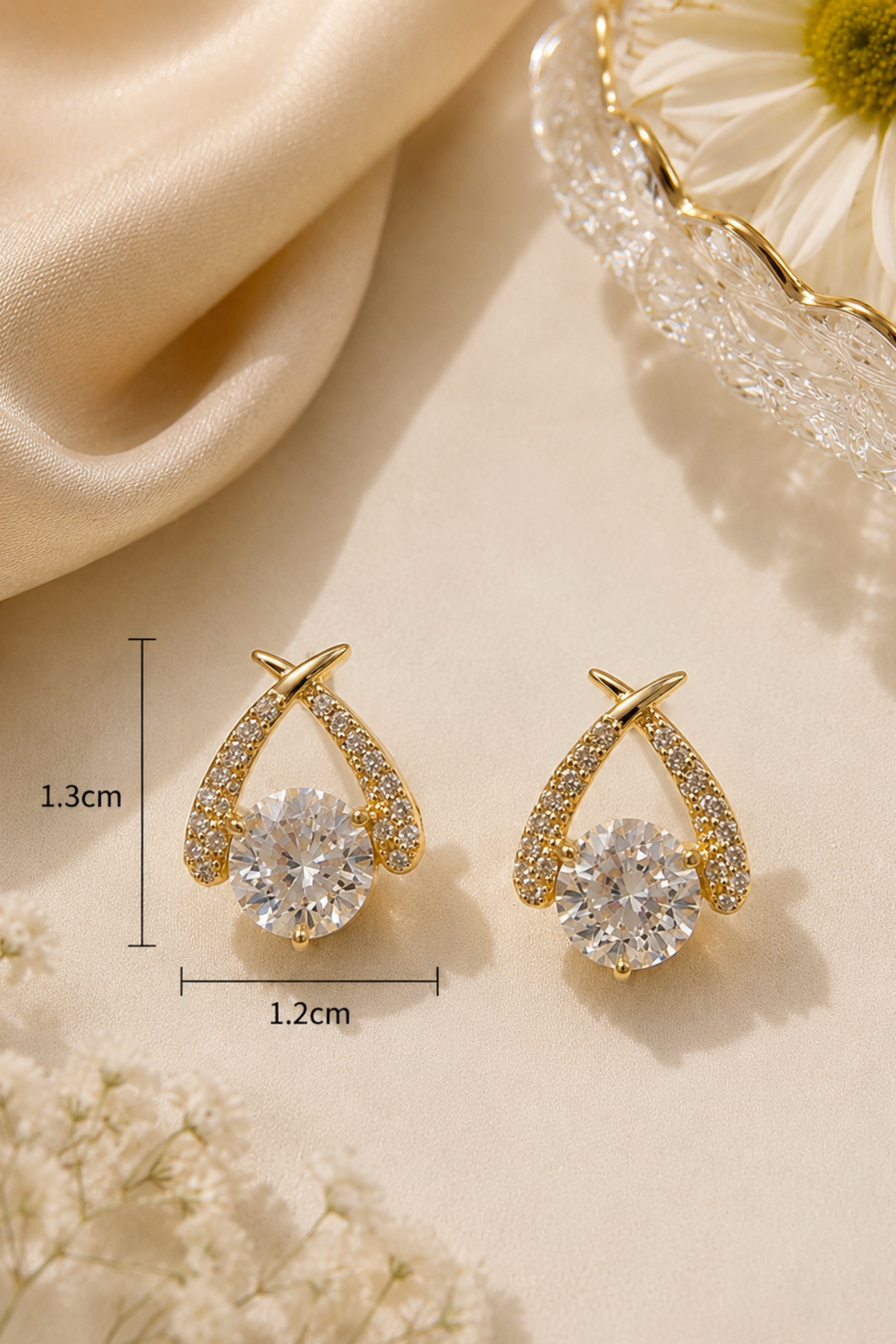 A pair of gold teardrop-shaped stud earrings with clear crystal pavé accents and a central round clear crystal, shown on a beige fabric background with decorative flowers and dimension labels indicating 1.3 cm height and 1.