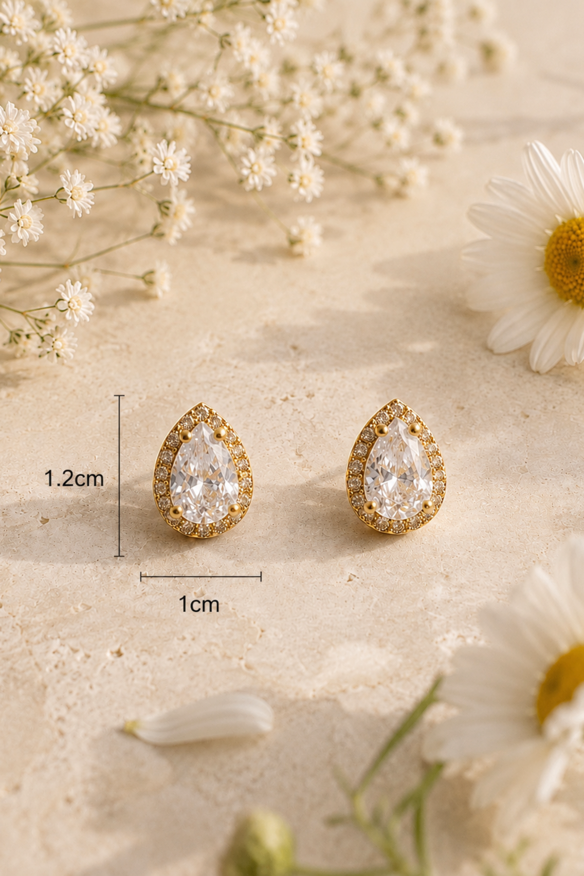 A pair of gold teardrop-shaped stud earrings with clear gemstones, displayed on a light beige textured surface with white flowers. A dimension overlay indicates the earrings are 1.