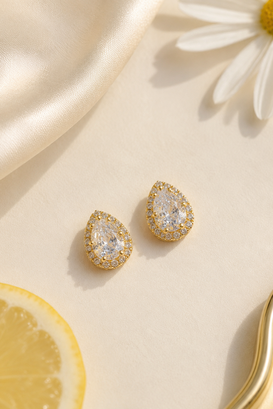 A pair of gold teardrop-shaped stud earrings with clear gemstones, styled on a cream surface with a satin fabric, a daisy flower, and a lemon slice.