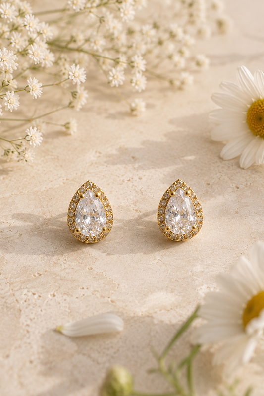 A pair of gold teardrop-shaped stud earrings with clear teardrop gemstones and small surrounding clear stones, styled on a light beige textured surface with white flowers.