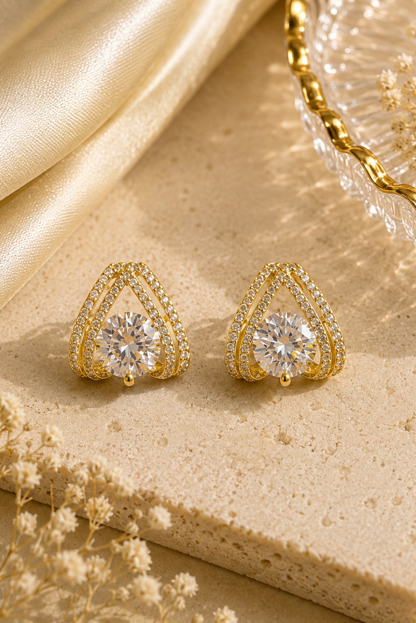 A pair of gold triangular stud earrings with a large clear center stone and small surrounding stones, displayed on a beige stone surface with gold fabric and dried flowers.
