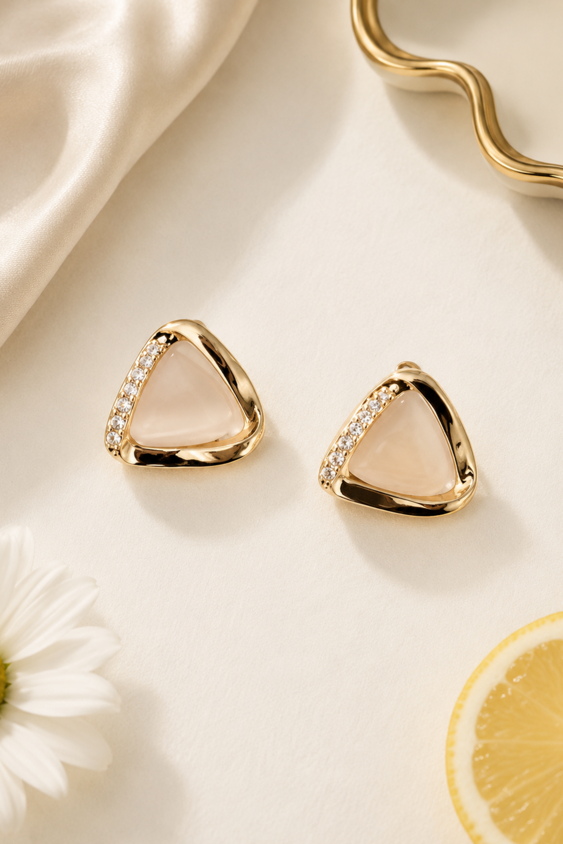 A pair of gold triangular stud earrings with a pale pink center and small clear stones along one side, styled on a cream surface with satin fabric, a daisy flower, and a lemon slice.