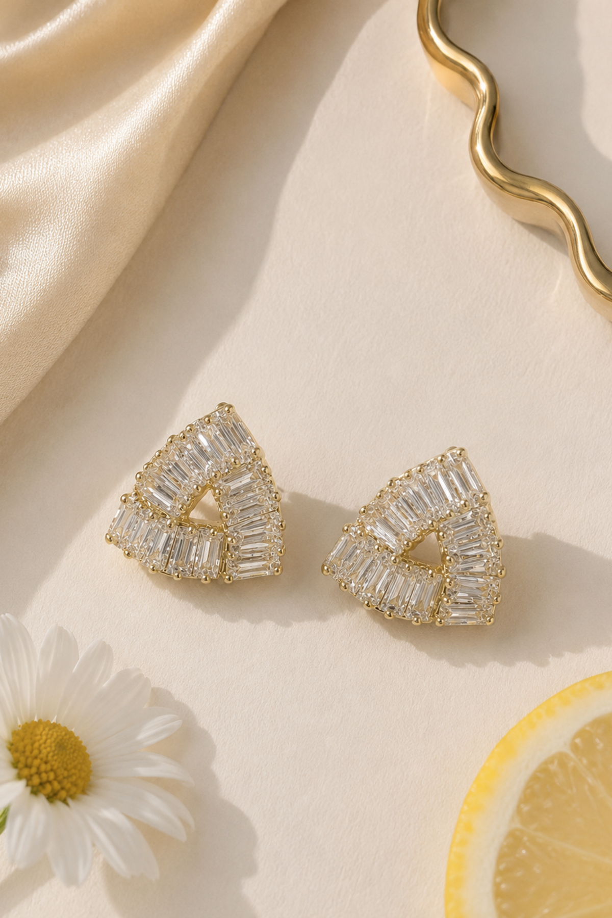 A pair of gold triangular stud earrings with clear baguette-cut stones, styled on a cream surface with a satin fabric, a daisy flower, and a lemon slice.
