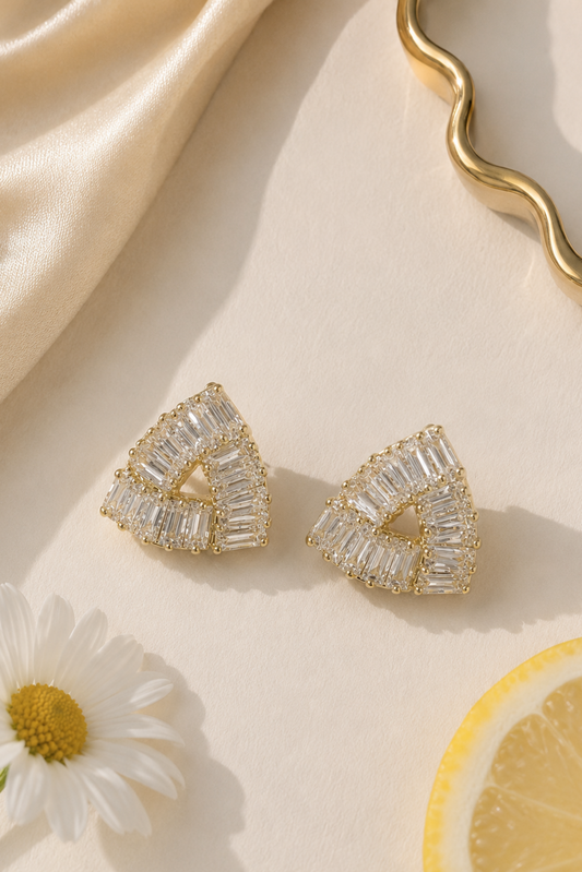 A pair of gold triangular stud earrings with clear baguette-cut stones, styled on a cream surface with a satin fabric, a daisy flower, and a lemon slice.